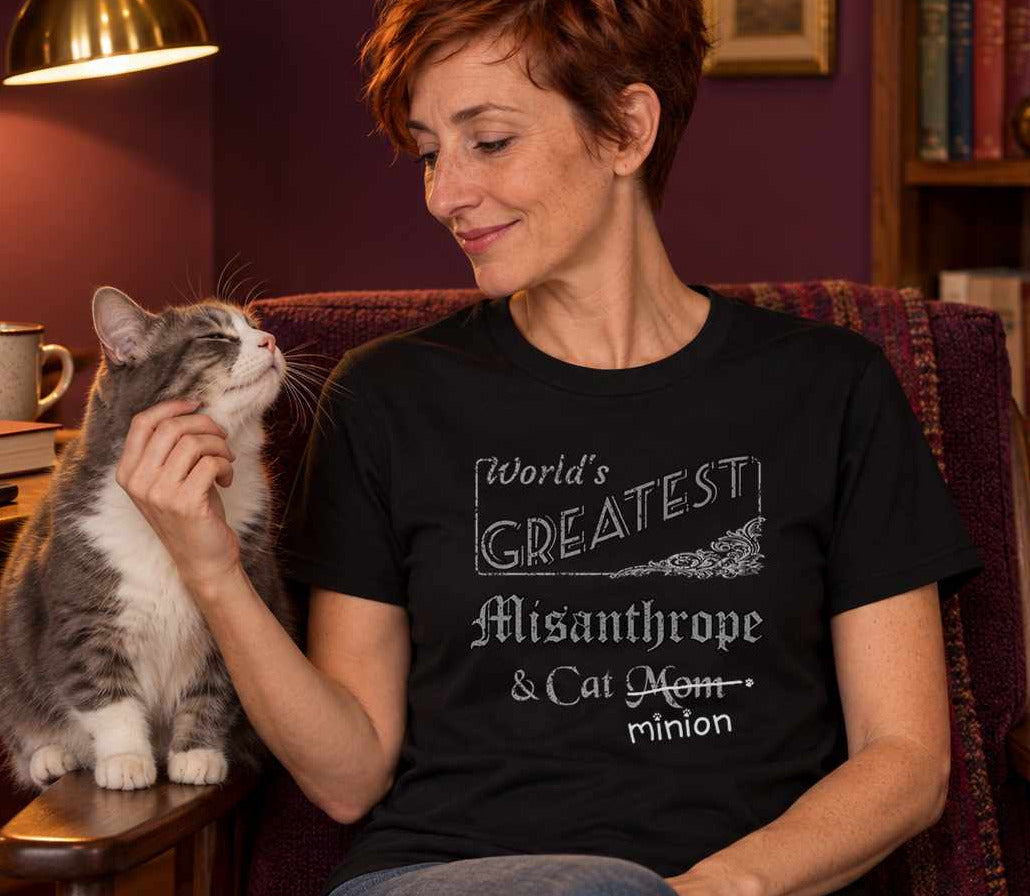 Woman scratching a grey-and-white cat’s cheek in a warmly lit plum reading nook, wearing a black crew-neck t-shirt that reads, “World’s Greatest Misanthrope & Cat Mom” with “Mom” crossed out and “minion” handwritten below with paw prints.