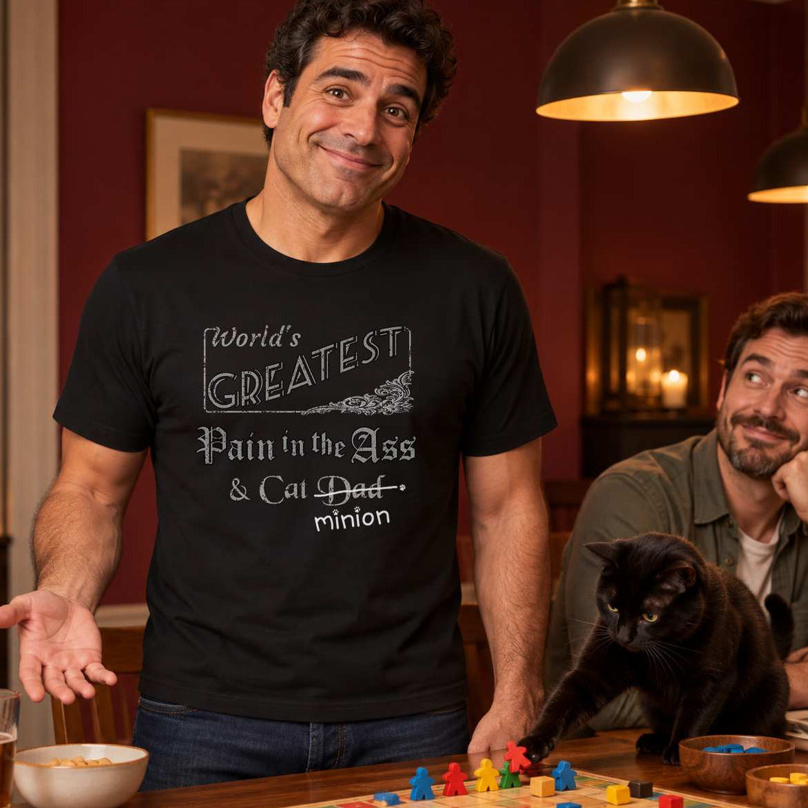 Man gesturing innocently while a black cat moves board-game pieces beside an amused friend, wearing a black crew-neck t-shirt that reads, “World’s Greatest Pain in the Ass & Cat Dad” with “Dad” crossed out and “minion” handwritten below with paw prints.