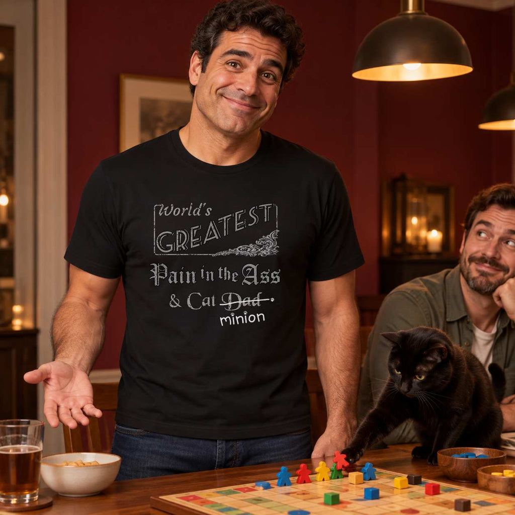 Man gesturing innocently while a black cat moves board-game pieces beside an amused friend, wearing a black crew-neck t-shirt that reads, “World’s Greatest Pain in the Ass & Cat Dad” with “Dad” crossed out and “minion” handwritten below with paw prints.