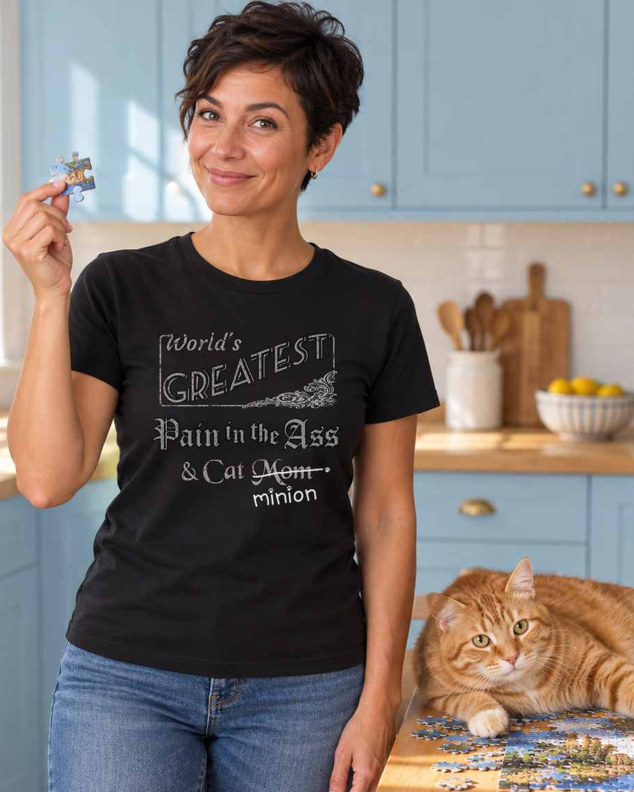 Woman holding a jigsaw piece while a ginger cat sprawls across the puzzle beside an amused companion, wearing a black crew-neck t-shirt that reads, “World’s Greatest Pain in the Ass & Cat Mom” with “Mom” crossed out and “minion” handwritten below with paw prints.