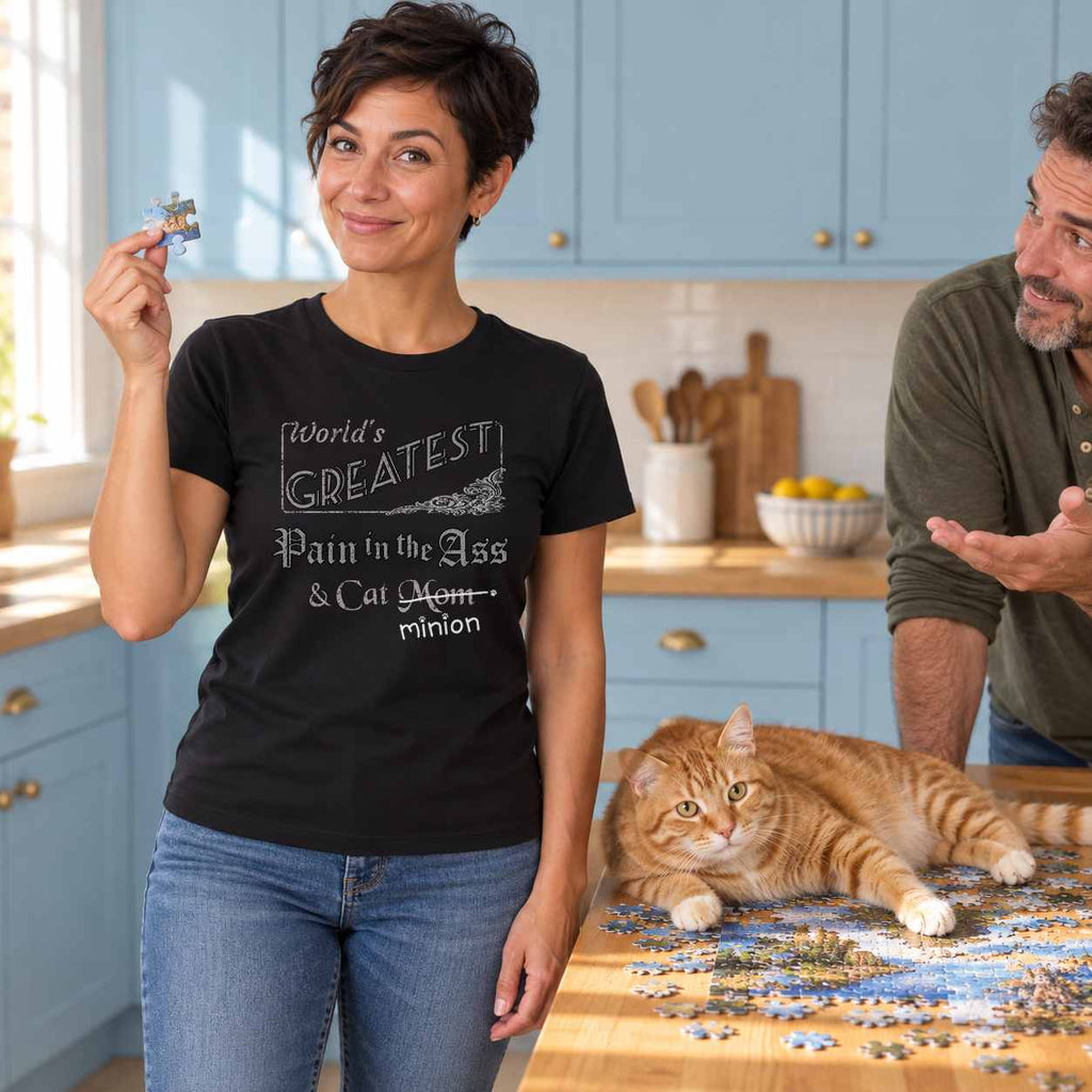 Woman holding a jigsaw piece while a ginger cat sprawls across the puzzle beside an amused companion, wearing a black crew-neck t-shirt that reads, “World’s Greatest Pain in the Ass & Cat Mom” with “Mom” crossed out and “minion” handwritten below with paw prints.