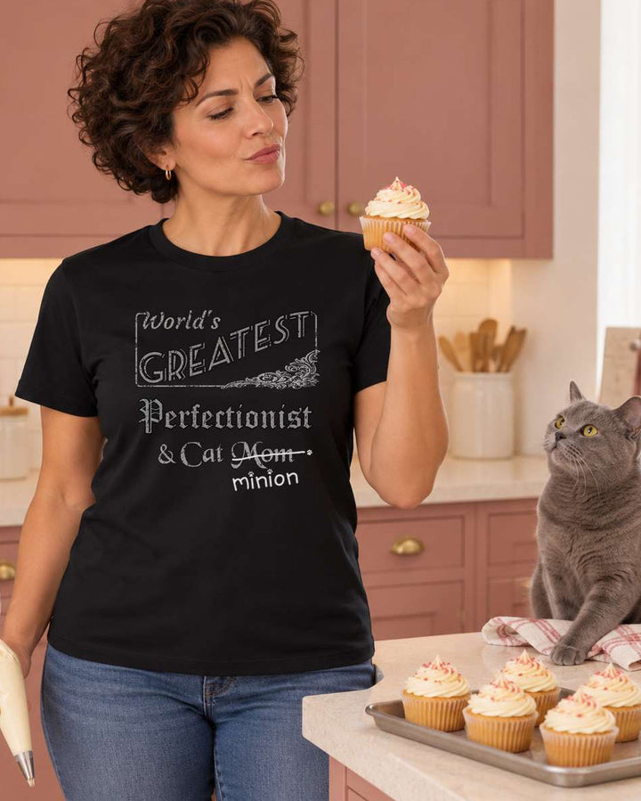 Woman inspecting a cupcake while holding a piping bag beside a watchful grey cat in a dusty-rose kitchen, wearing a black crew-neck t-shirt that reads, “World’s Greatest Perfectionist & Cat Mom” with “Mom” crossed out and “minion” handwritten below with paw prints.