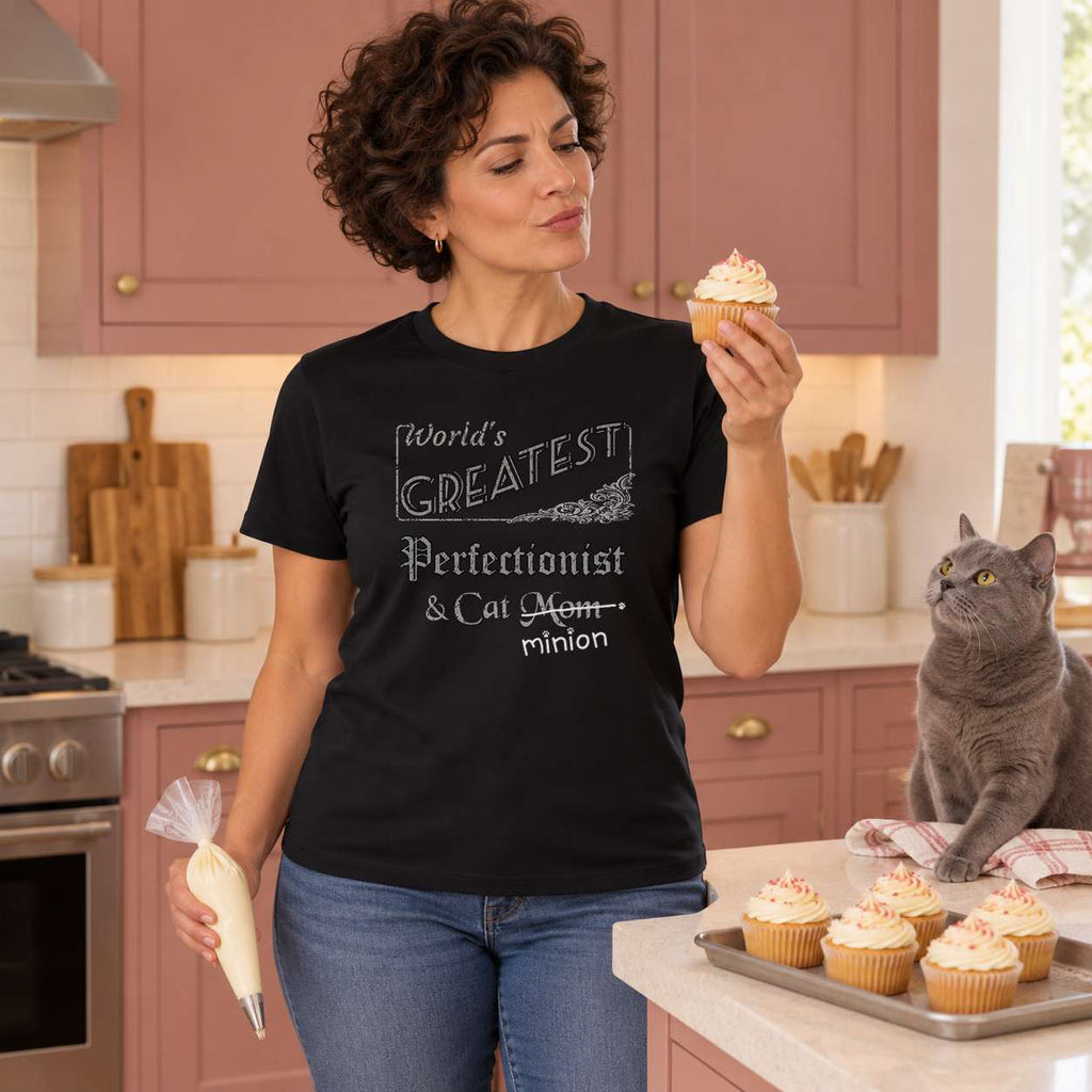 Woman inspecting a cupcake while holding a piping bag beside a watchful grey cat in a dusty-rose kitchen, wearing a black crew-neck t-shirt that reads, “World’s Greatest Perfectionist & Cat Mom” with “Mom” crossed out and “minion” handwritten below with paw prints.