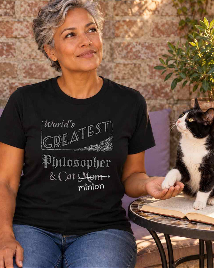 Woman holding a tuxedo cat’s paw beside an open book in a sheltered garden reading spot, wearing a black crew-neck t-shirt that reads, “World’s Greatest Philosopher & Cat Mom” with “Mom” crossed out and “minion” handwritten below with paw prints.