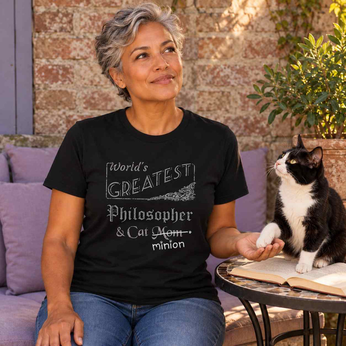 Woman holding a tuxedo cat’s paw beside an open book in a sheltered garden reading spot, wearing a black crew-neck t-shirt that reads, “World’s Greatest Philosopher & Cat Mom” with “Mom” crossed out and “minion” handwritten below with paw prints.
