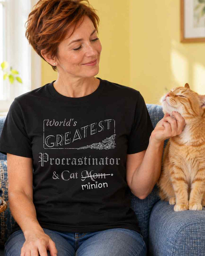 Woman scratching a ginger cat’s chin beside a basket of unfolded laundry in a sunny room, wearing a black crew-neck t-shirt that reads, “World’s Greatest Procrastinator & Cat Mom” with “Mom” crossed out and “minion” handwritten below with paw prints.