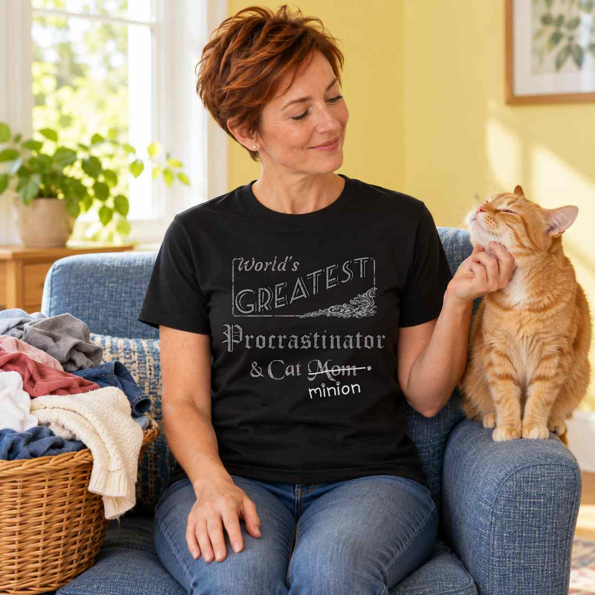 Woman scratching a ginger cat’s chin beside a basket of unfolded laundry in a sunny room, wearing a black crew-neck t-shirt that reads, “World’s Greatest Procrastinator & Cat Mom” with “Mom” crossed out and “minion” handwritten below with paw prints.