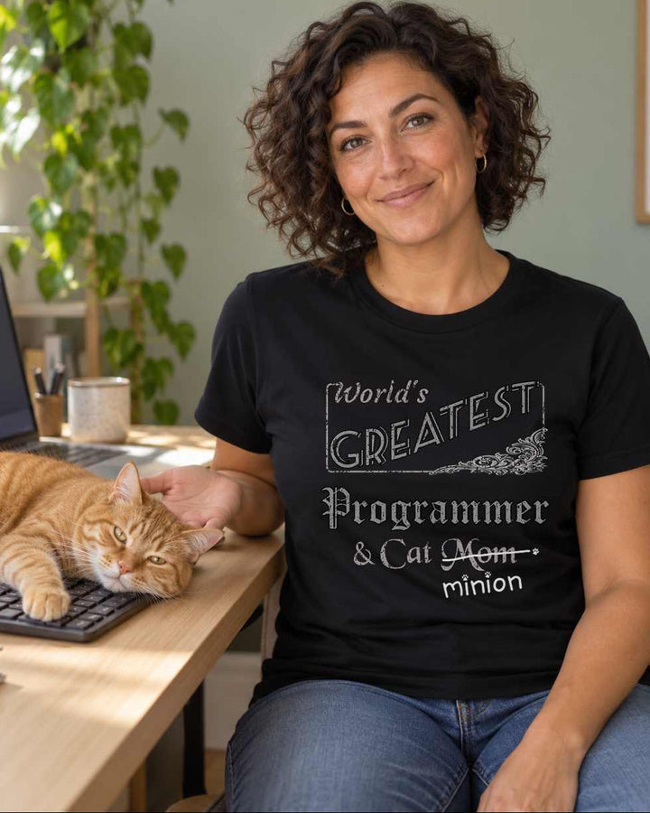 Woman presenting an orange tabby sprawled across a keyboard beside her laptop, wearing a black crew-neck t-shirt that reads, “World’s Greatest Programmer & Cat Mom” with “Mom” crossed out and “minion” handwritten below with paw prints.