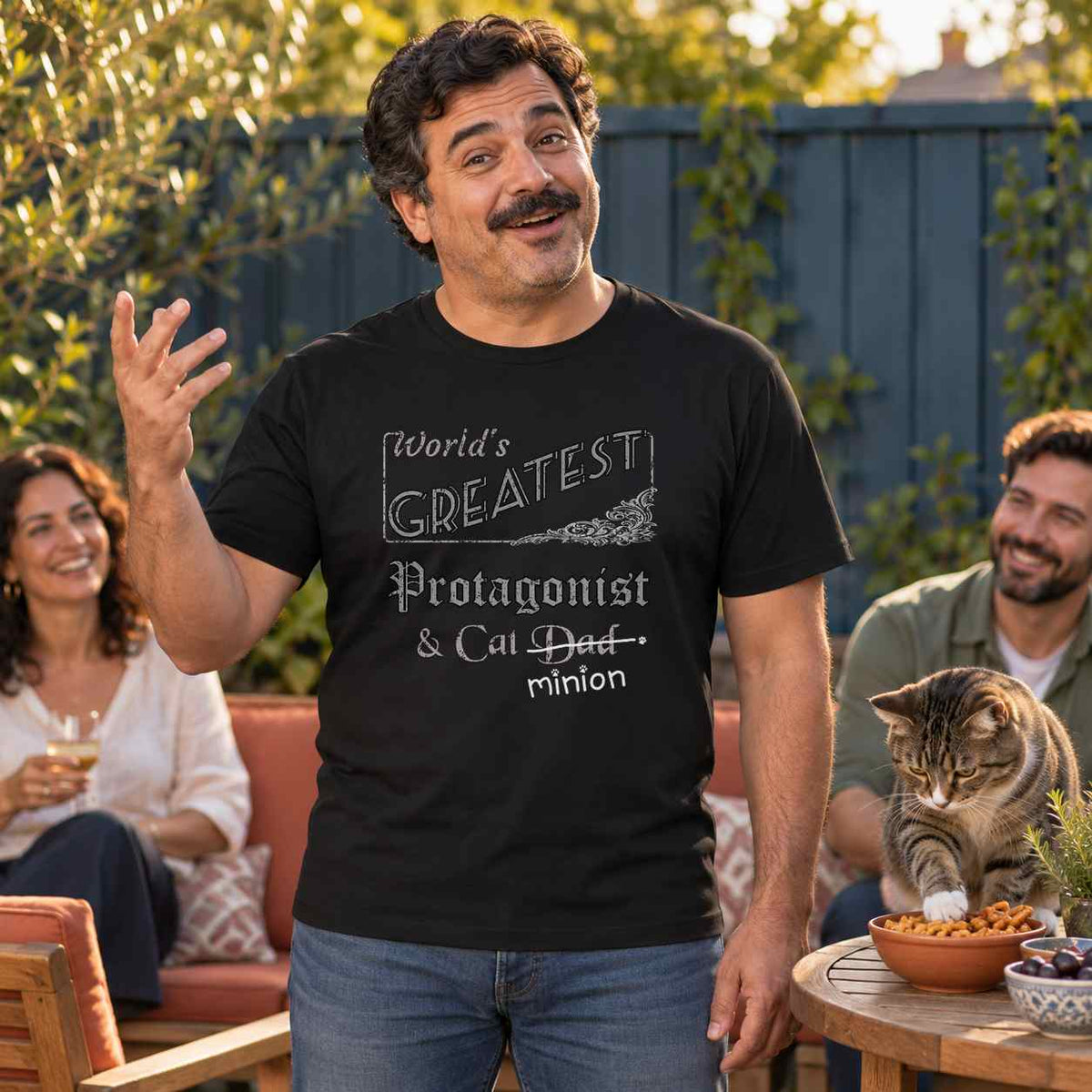 Man gesturing while telling a story at a backyard gathering as a tabby cat reaches into a snack bowl, wearing a black crew-neck t-shirt that reads, “World’s Greatest Protagonist & Cat Dad” with “Dad” crossed out and “minion” handwritten below with paw prints.