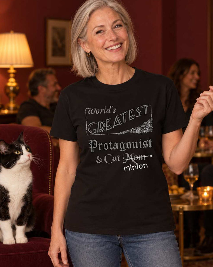 Woman telling a story in a warmly lit burgundy living room beside a tuxedo cat sitting in an armchair, wearing a black crew-neck t-shirt that reads, “World’s Greatest Protagonist & Cat Mom” with “Mom” crossed out and “minion” handwritten below with paw prints.