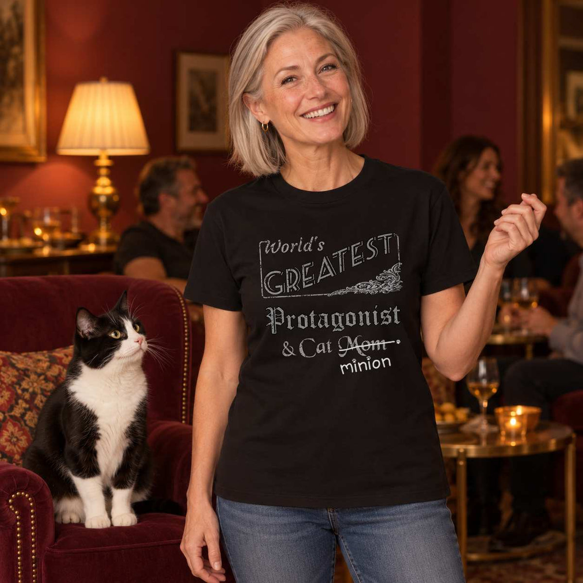 Woman telling a story in a warmly lit burgundy living room beside a tuxedo cat sitting in an armchair, wearing a black crew-neck t-shirt that reads, “World’s Greatest Protagonist & Cat Mom” with “Mom” crossed out and “minion” handwritten below with paw prints.
