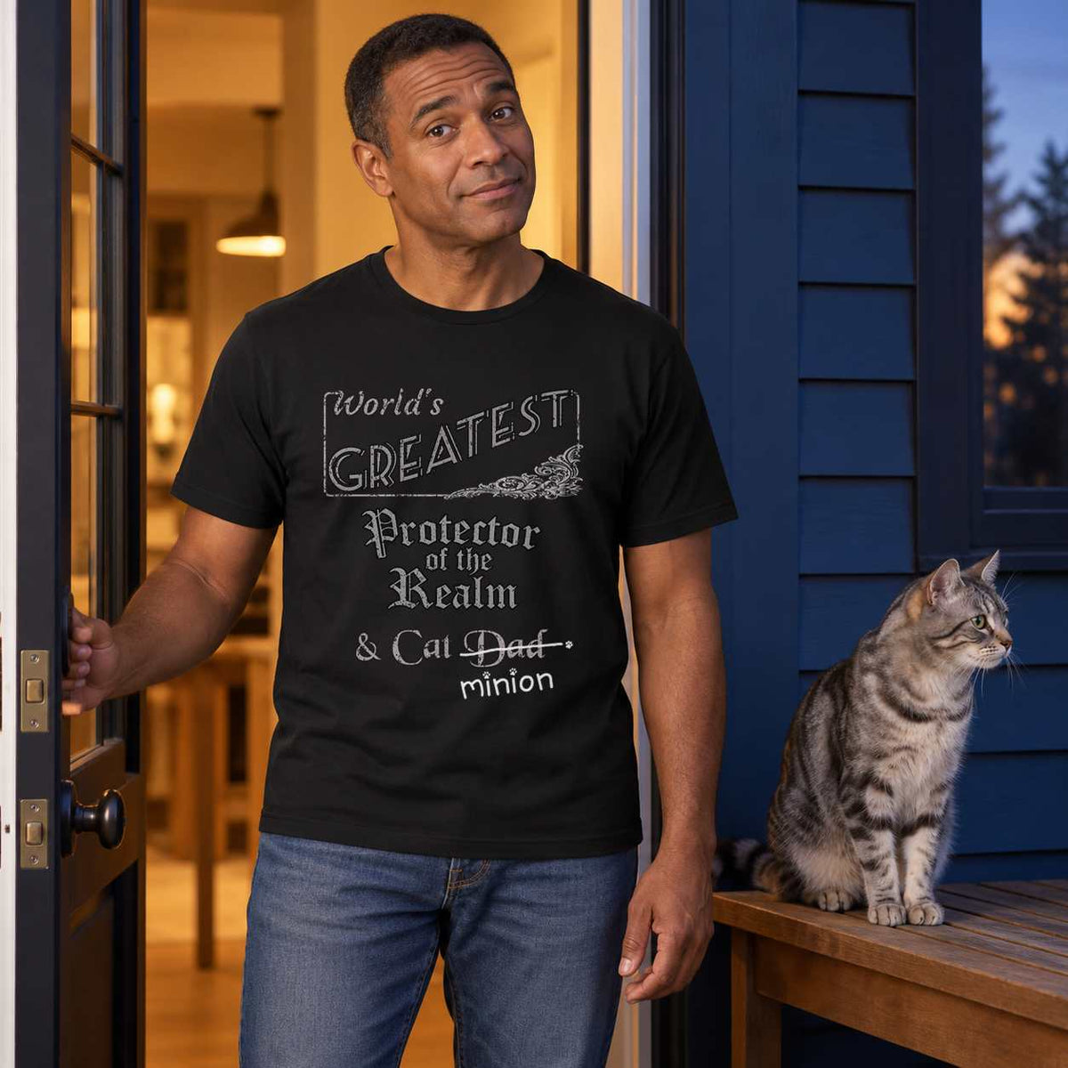 Man holding a back door open at dusk beside a silver tabby cat sitting on a bench, wearing a black crew-neck t-shirt that reads, “World’s Greatest Protector of the Realm & Cat Dad” with “Dad” crossed out and “minion” handwritten below with paw prints.