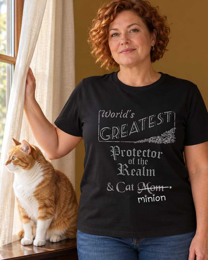 Woman holding a curtain aside while a ginger and white cat surveys outdoors from a wooden console, wearing a black crew-neck t-shirt that reads, “World’s Greatest Protector of the Realm & Cat Mom” with “Mom” crossed out and “minion” handwritten below with paw prints.