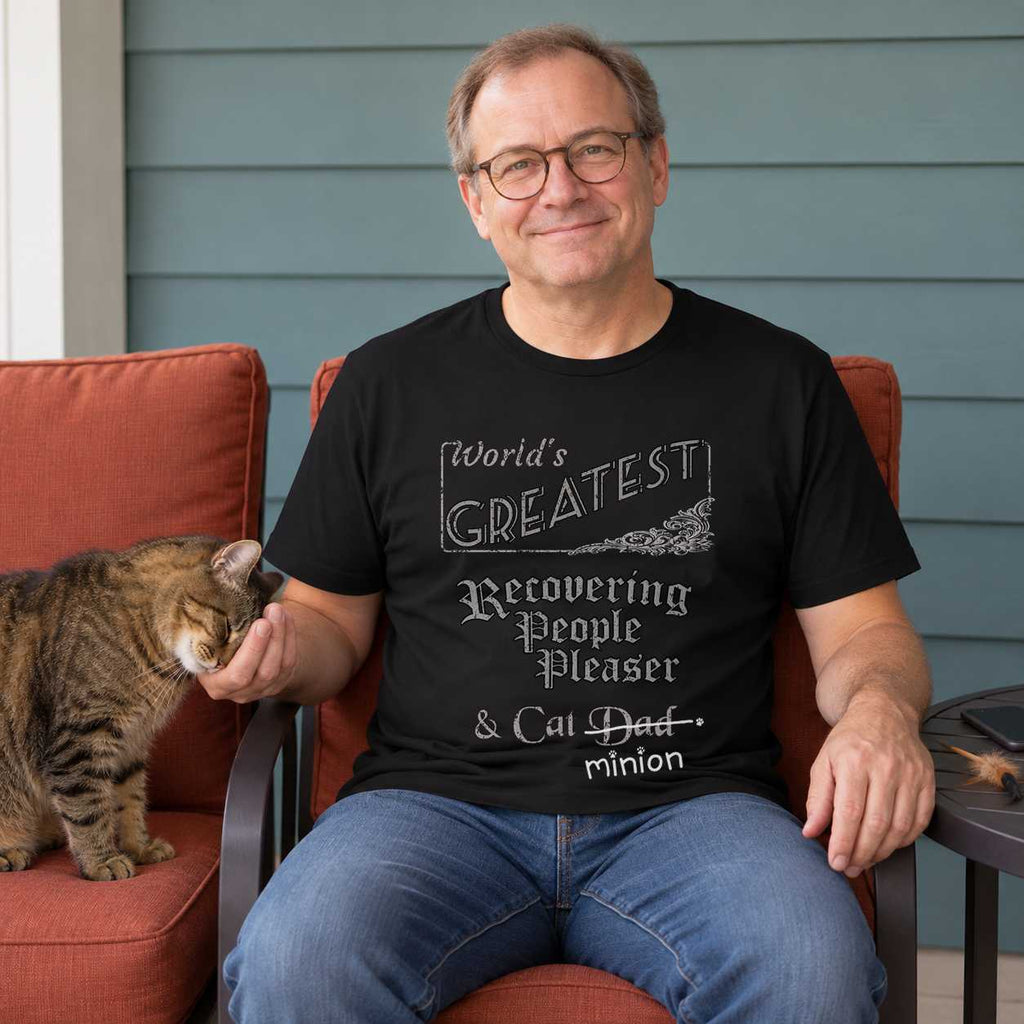 Man relaxing on a teal patio as a tabby cat presses its head into his hand, with his phone and feather toy set aside, wearing a black crew-neck t-shirt that reads, “World’s Greatest Recovering People Pleaser & Cat Dad” with “Dad” crossed out and “minion” handwritten below with paw prints.