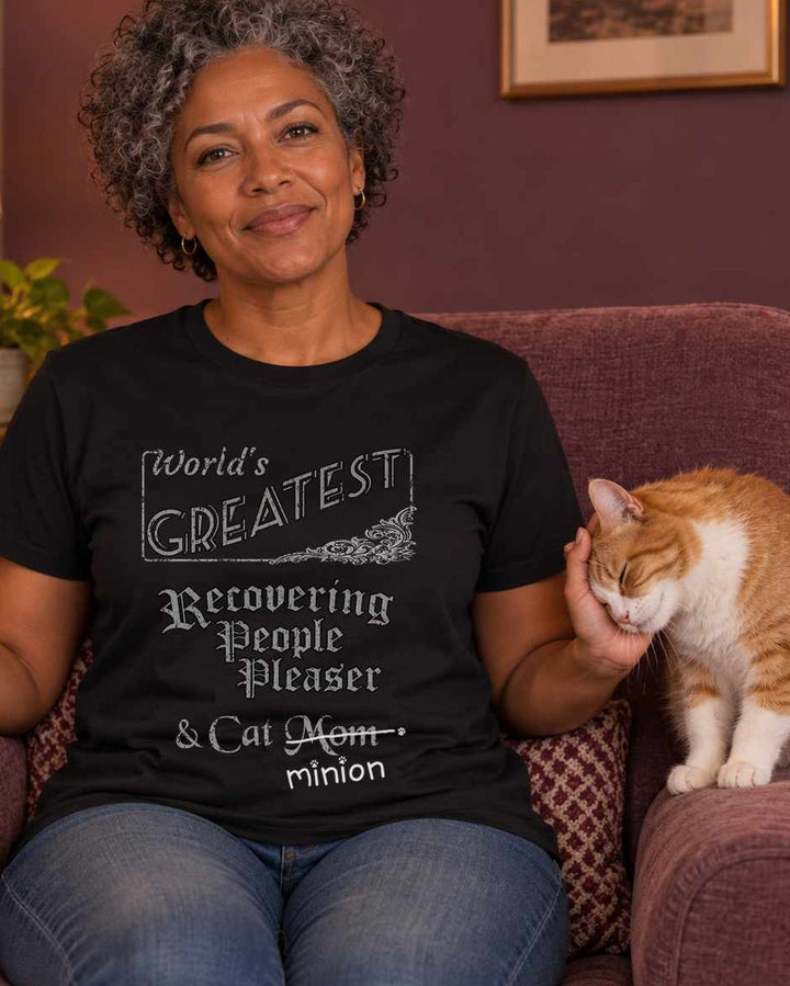 Woman holding a mug in a warmly lit plum living room as a ginger and white cat presses its head into her free hand, wearing a black crew-neck t-shirt that reads, “World’s Greatest Recovering People Pleaser & Cat Mom” with “Mom” crossed out and “minion” handwritten below with paw prints.