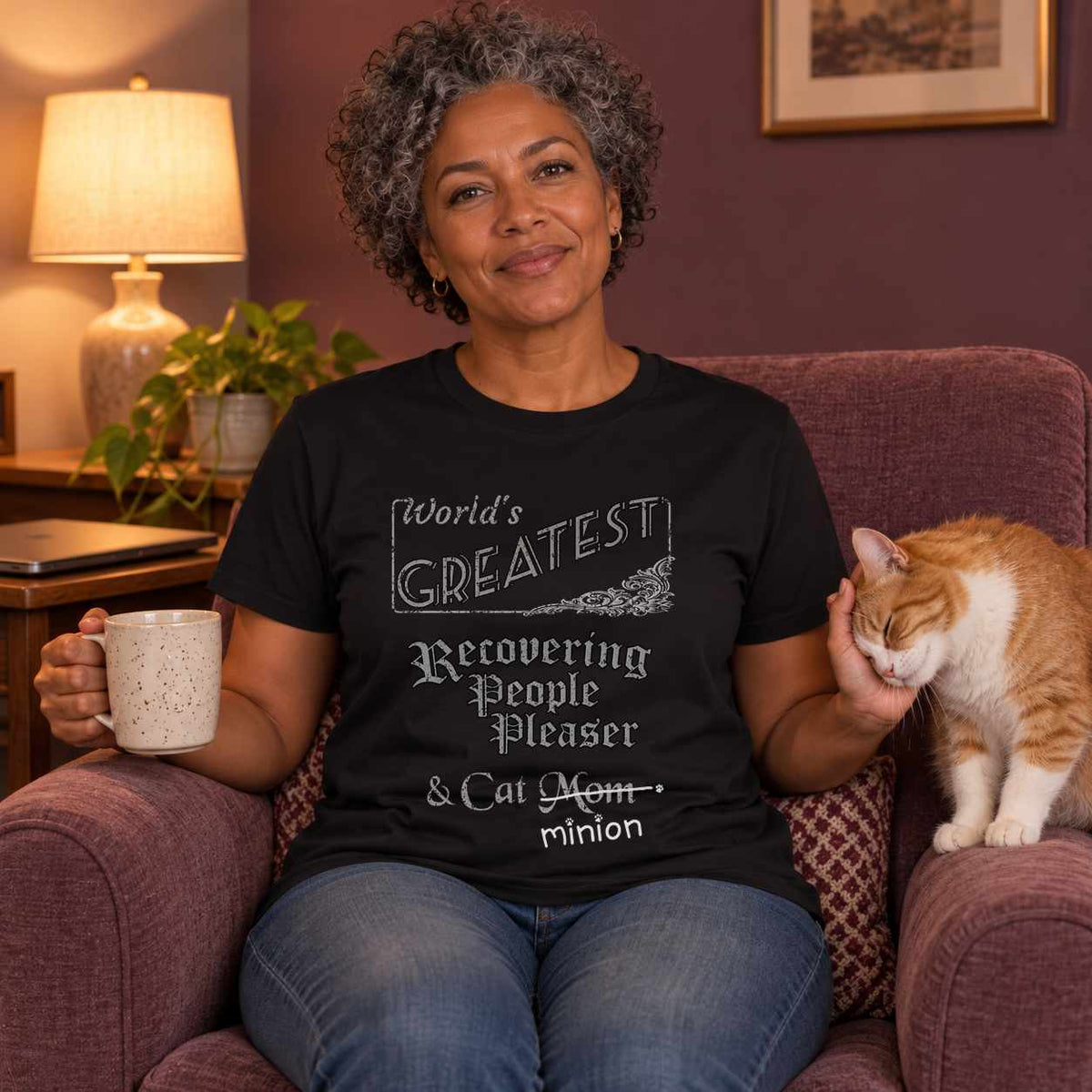 Woman holding a mug in a warmly lit plum living room as a ginger and white cat presses its head into her free hand, wearing a black crew-neck t-shirt that reads, “World’s Greatest Recovering People Pleaser & Cat Mom” with “Mom” crossed out and “minion” handwritten below with paw prints.