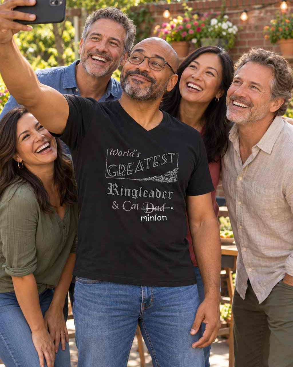 Man taking a selfie in the centre of a group of friends, wearing a black V-neck T-shirt reading “World’s Greatest Ringleader & Cat Dad.” “Dad” is crossed out, with “minion” handwritten beneath it and surrounded by cat paw prints.