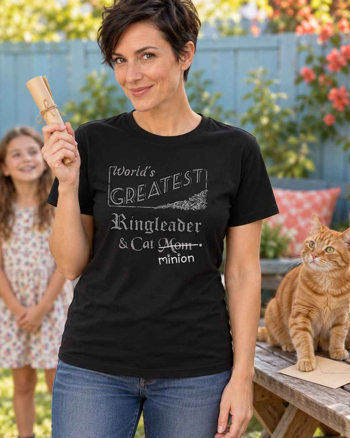 Woman holding a rolled treasure-hunt clue with two children waiting and a ginger cat pinning an envelope on a picnic table, wearing a black crew-neck t-shirt that reads, “World’s Greatest Ringleader & Cat Mom” with “Mom” crossed out and “minion” handwritten below with paw prints.