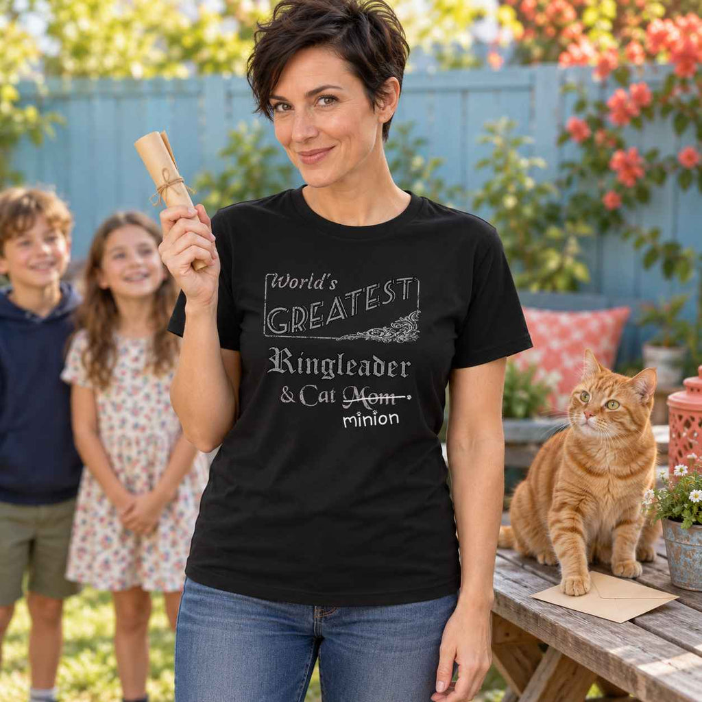 Woman holding a rolled treasure-hunt clue with two children waiting and a ginger cat pinning an envelope on a picnic table, wearing a black crew-neck t-shirt that reads, “World’s Greatest Ringleader & Cat Mom” with “Mom” crossed out and “minion” handwritten below with paw prints.