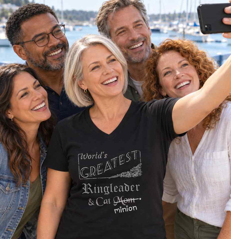 Woman taking a selfie in the centre of a group of friends, wearing a black V-neck T-shirt reading “World’s Greatest Ringleader & Cat Mom.” “Mom” is crossed out, with “minion” handwritten beneath it and surrounded by cat paw prints.