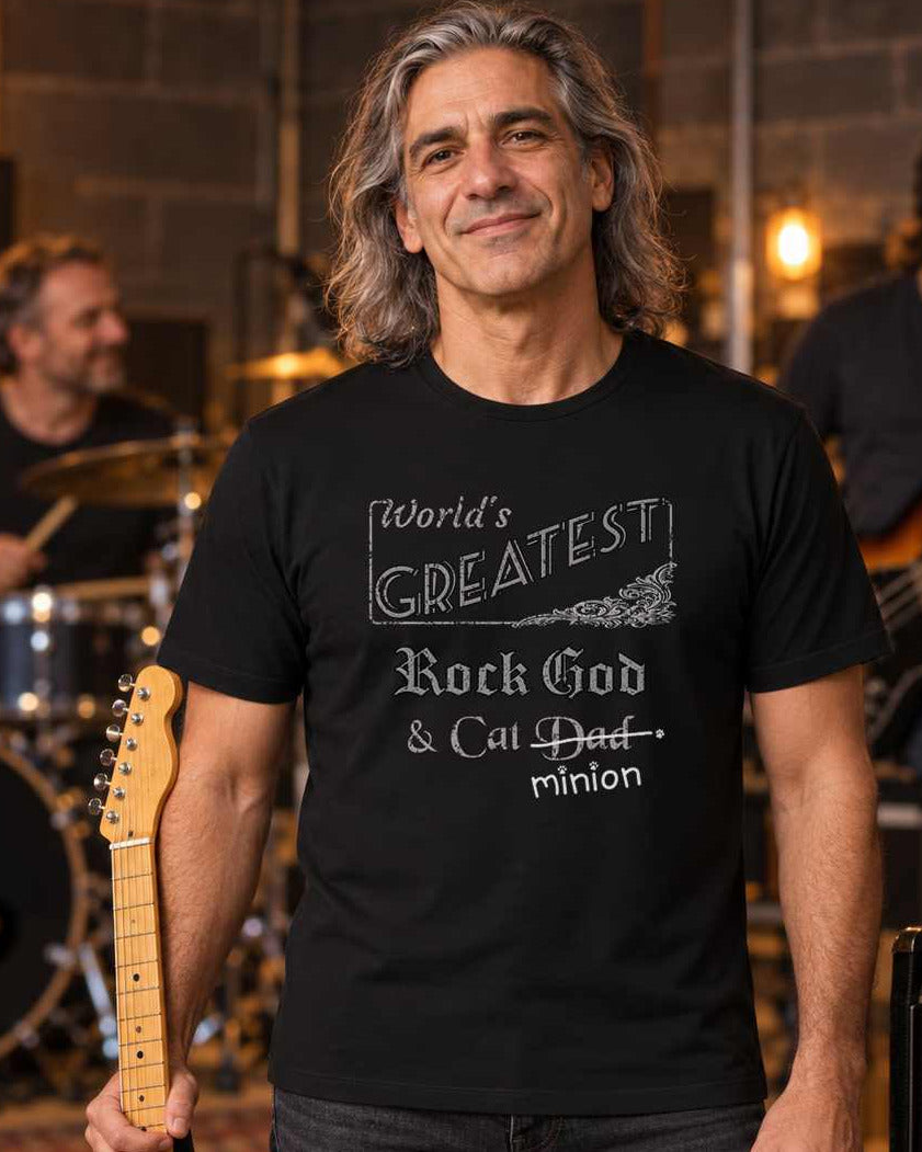 Man holding an electric guitar beside a tabby cat on an amplifier, with bandmates rehearsing behind him, wearing a black crew-neck t-shirt that reads, “World’s Greatest Rock God & Cat Dad” with “Dad” crossed out and “minion” handwritten below with paw prints.