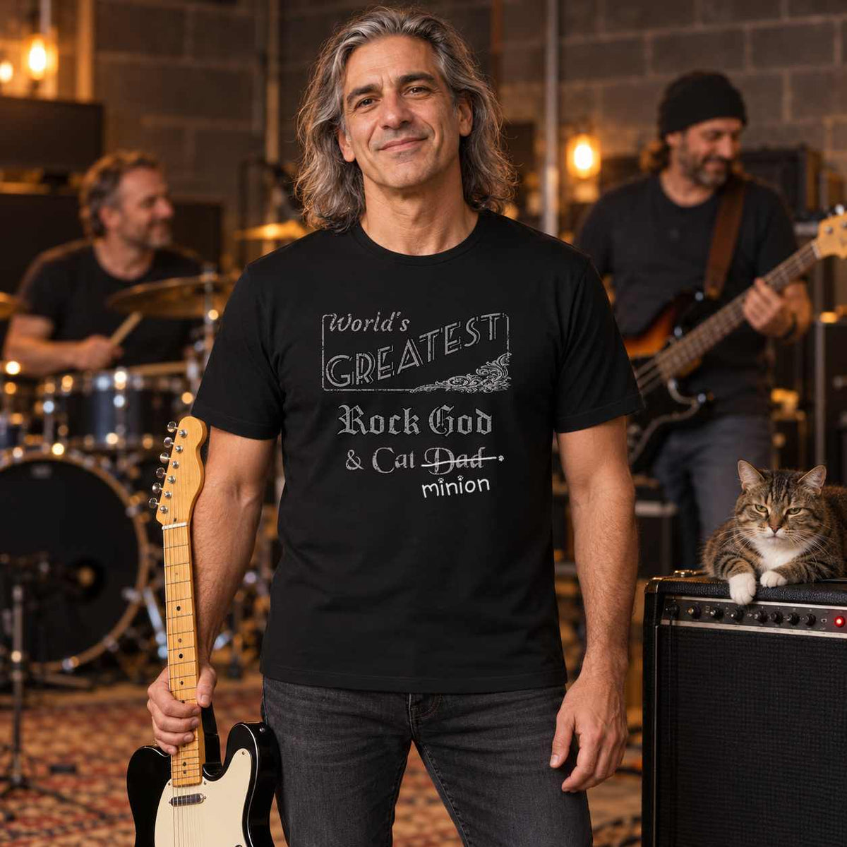 Man holding an electric guitar beside a tabby cat on an amplifier, with bandmates rehearsing behind him, wearing a black crew-neck t-shirt that reads, “World’s Greatest Rock God & Cat Dad” with “Dad” crossed out and “minion” handwritten below with paw prints.