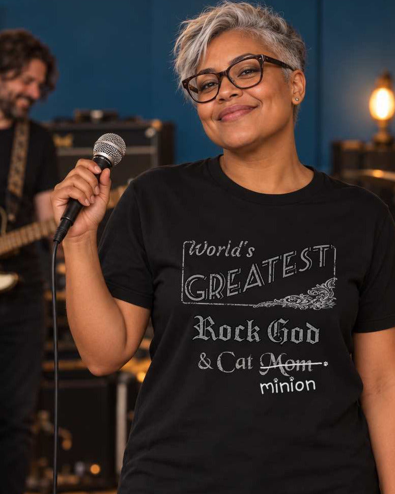 Woman holding a microphone beside a ginger and white cat on a guitar case, with bandmates rehearsing behind her, wearing a black crew-neck t-shirt that reads, “World’s Greatest Rock God & Cat Mom” with “Mom” crossed out and “minion” handwritten below with paw prints.