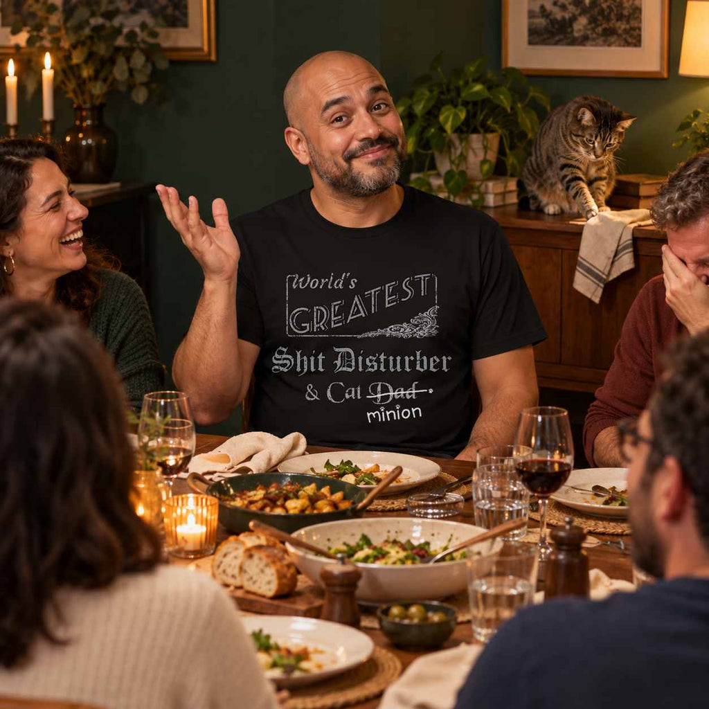 Man gesturing innocently at a dinner gathering as friends laugh and a tabby cat nudges a napkin off a sideboard, wearing a black crew-neck t-shirt that reads, “World’s Greatest Shit Disturber & Cat Dad” with “Dad” crossed out and “minion” handwritten below with paw prints.