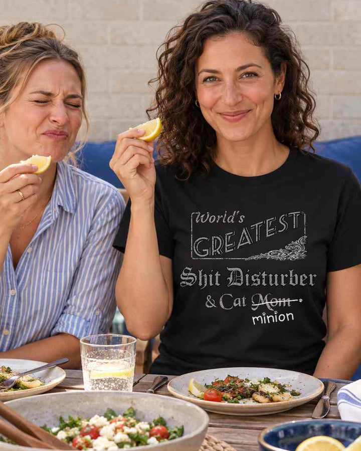 Woman holding a lemon wedge at a lunch gathering beside a friend making a sour face while a tuxedo cat tugs a napkin, wearing a black crew-neck t-shirt that reads, “World’s Greatest Shit Disturber & Cat Mom” with “Mom” crossed out and “minion” handwritten below with paw prints.