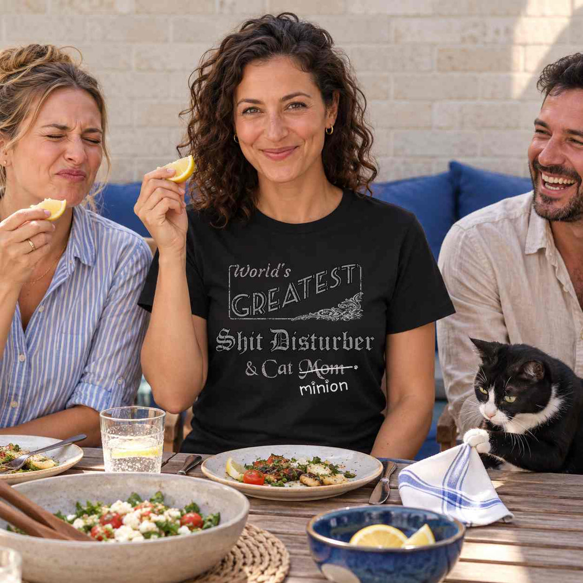 Woman holding a lemon wedge at a lunch gathering beside a friend making a sour face while a tuxedo cat tugs a napkin, wearing a black crew-neck t-shirt that reads, “World’s Greatest Shit Disturber & Cat Mom” with “Mom” crossed out and “minion” handwritten below with paw prints.