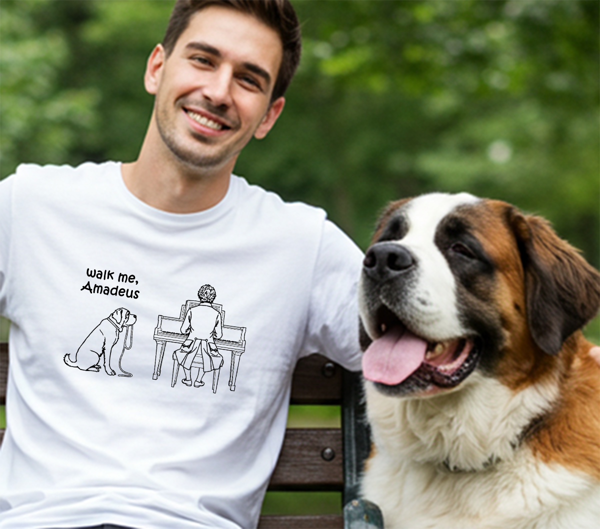 A smiling man wearing a white graphic T-shirt sits on a park bench next to a large Saint Bernard dog. The shirt has a black line drawing of the dog sitting next to a person playing a piano, with the text 'walk me, Amadeus'.