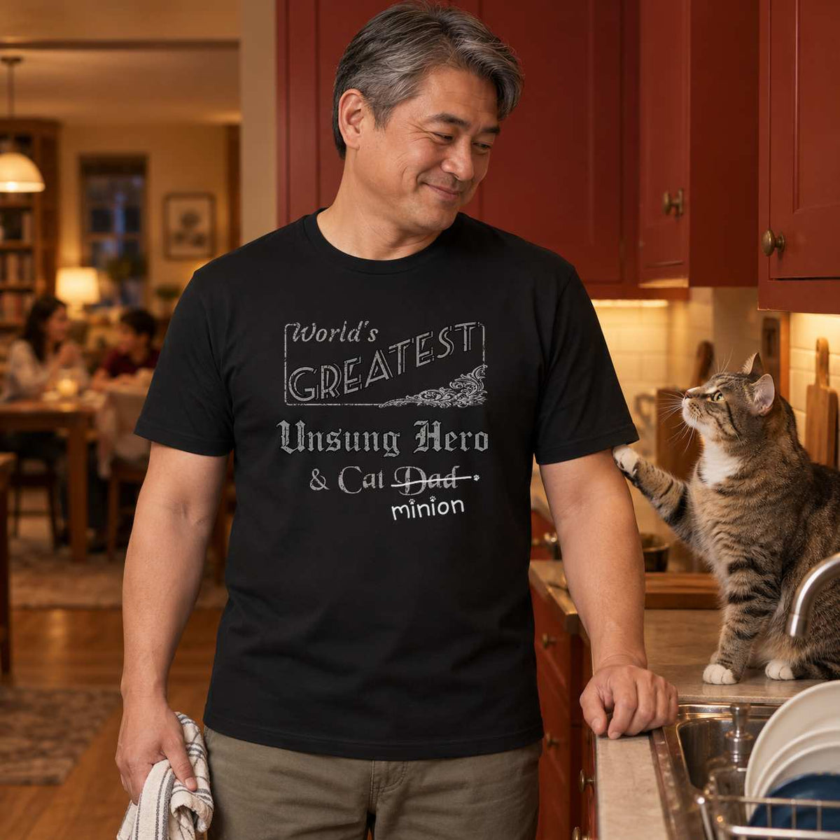 Man finishing the washing-up while a tabby cat taps his arm, wearing a black crew-neck t-shirt that reads, “World’s Greatest Unsung Hero & Cat Dad” with “Dad” crossed out and “minion” handwritten below with paw prints.