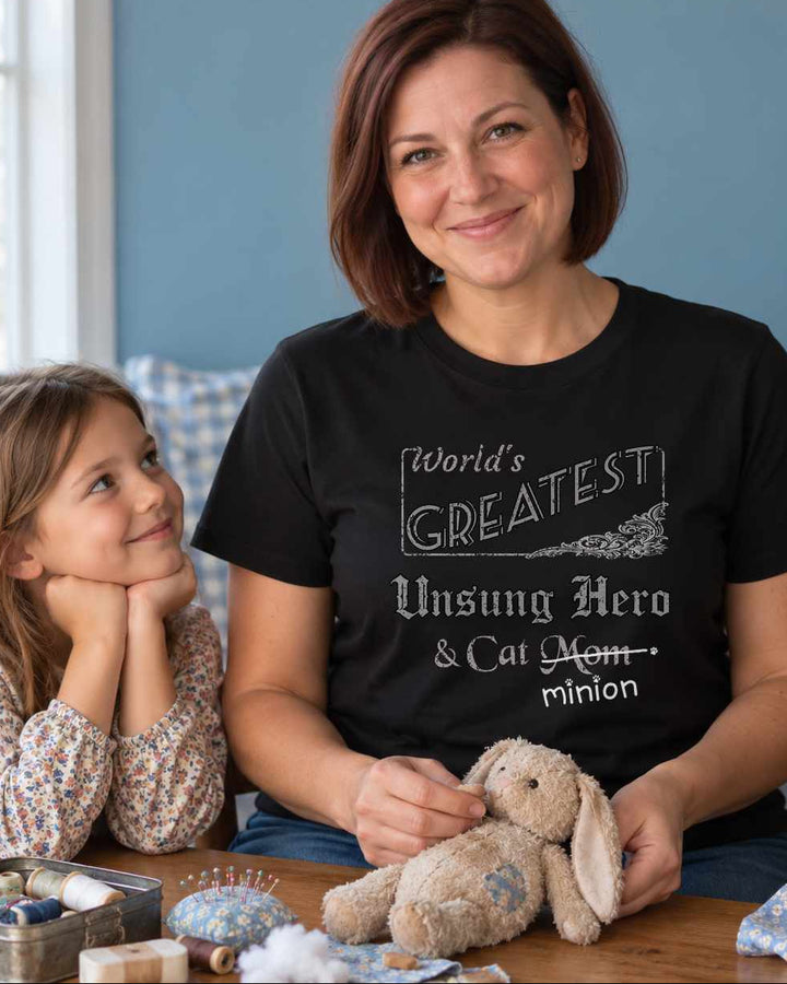 Woman repairing a stuffed rabbit beside a child while a cat holds spare fabric under its paw, wearing a black crew-neck t-shirt that reads, “World’s Greatest Unsung Hero & Cat Mom” with “Mom” crossed out and “minion” handwritten below with paw prints.