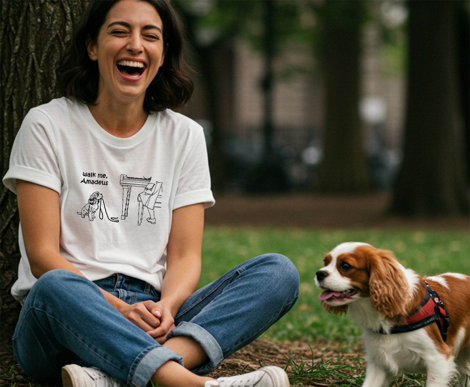 A woman sitting on the grass in a park next to a Cavalier King Charles Spaniel dog, wearing a white graphic t-shirt with a black line art illustration of Napoleon and the text 'Walk me, Amadeus'.