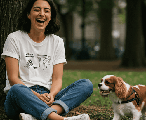 A woman sitting on the grass in a park next to a Cavalier King Charles Spaniel dog, wearing a white graphic t-shirt with a bl