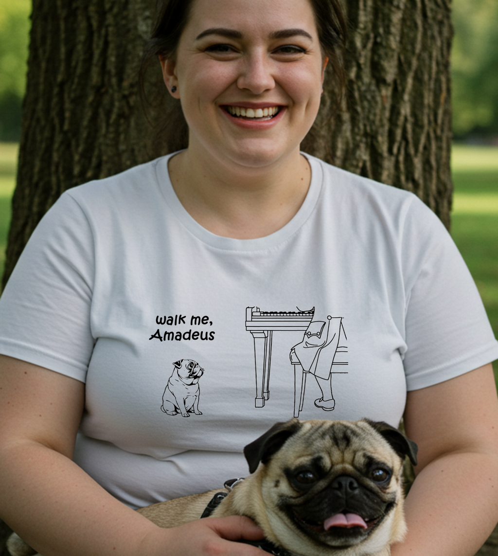 A woman smiling outdoors in front of a tree, wearing a white graphic T-shirt with a black line drawing of a pug and a piano, plus the text 'walk me, Amadeus.' She is holding a pug in a park-like setting.