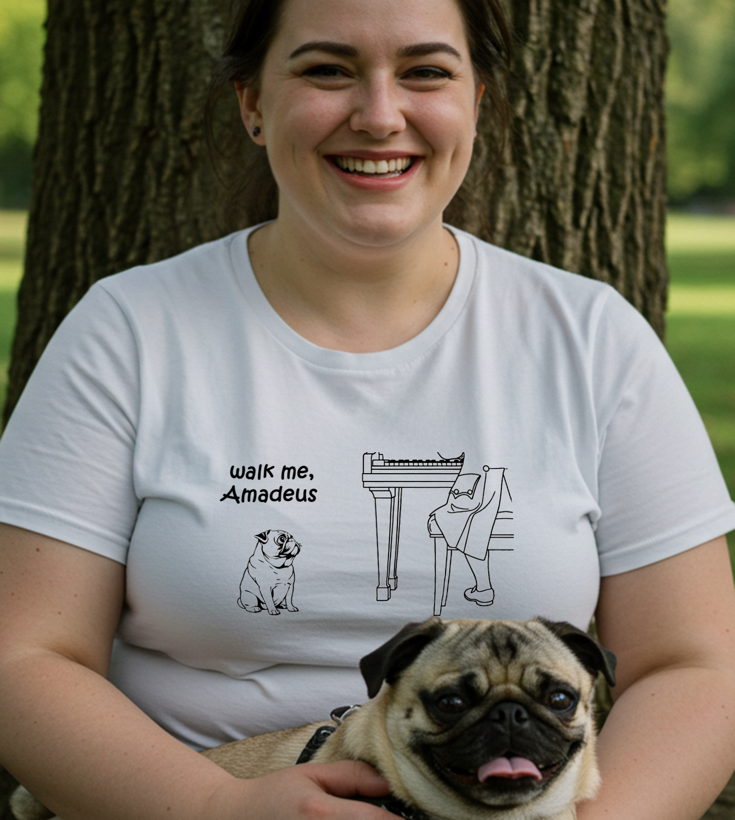 A woman smiling outdoors in front of a tree, wearing a white graphic T-shirt with a black line drawing of a pug and a piano, plus the text 'walk me, Amadeus.' She is holding a pug in a park-like setting.